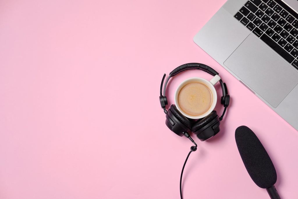 Flat lay of a podcast recording setup with headphones, a microphone, a cup of coffee, and a laptop on a pink background.